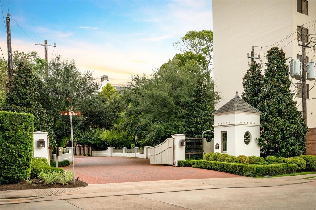 Landscaped community entrance with white walls, a small entry building, trees, and brick paving.